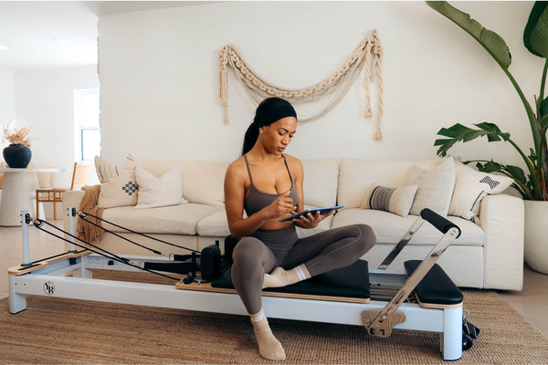 White Pilates Reformer Rental in Boho Living Room - Woman Using Tablet | Your Reformer